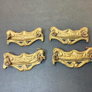 May include: Four antique brass drawer pulls with ornate, scroll-like designs. Each pull features a decorative face with a central cutout and small ring handles. The brass has a warm, aged patina, suggesting vintage or antique origins.
