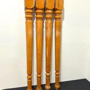 May include: Four matching light brown wooden furniture legs with a turned spindle design. Each leg features a square top block with pre-drilled holes for mounting, followed by a series of decorative rounded rings and a tapered cylindrical body.