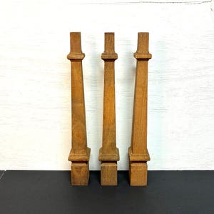 May include: Three wooden balusters with a warm, medium-brown finish. Each baluster has a rectangular base, a long, slender body, and a decorative top. They are arranged against a white and black background.