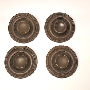 May include: Four antique bronze-colored drawer pulls with ring handles. Each pull has a circular design with a raised center and a dotted outer edge. The pulls are arranged in a square formation against a white background.