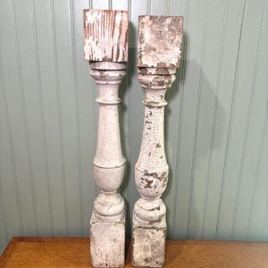 May include: Two tall, vertical wooden balusters featuring a distressed white painted finish with visible wood grain and chipped paint. Each piece has a square base, a central turned spindle section, and a square block at the top end.