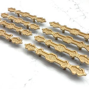 May include: A collection of gold-colored cabinet pulls with an ornate, vintage design. The pulls are arranged on a white marble surface, showcasing their decorative details and overall aesthetic. The hardware is likely for furniture.