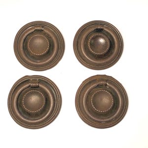 May include: A set of four circular bronze-toned metal drawer pulls. Each pull features a round backplate with a beaded edge and a central circular knob surrounded by a hinged ring handle that rests within the recessed area of the plate.