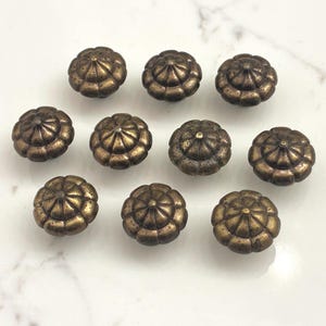 May include: Ten antique bronze-colored cabinet knobs with a detailed, rounded design. The knobs are arranged on a white surface, showcasing their intricate, floral-like pattern. These decorative knobs are ideal for furniture restoration or adding a vintage touch.