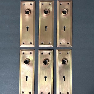 May include: Six antique brass door plates. Each rectangular plate features a keyhole, a circular opening for a doorknob, and screw holes. The plates have a warm, aged brass finish, suggesting vintage hardware.