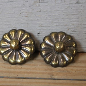 May include: Two antique brass flower-shaped cabinet knobs. Each knob features a central, rounded stud surrounded by a scalloped, petal-like design. The knobs have a warm, aged patina, suggesting vintage hardware.