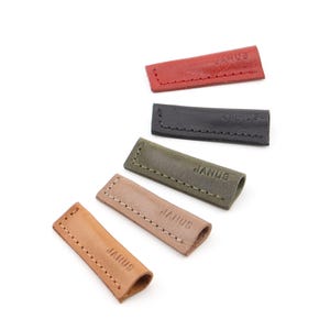 May include: A selection of leather pen holders in shades of red, black, olive green, tan, and light brown. Each holder is rectangular with rounded edges and stitched detailing. The word "JANUS" is embossed on each holder.