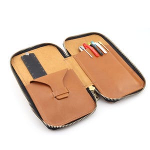May include: Open brown leather pencil case with a black zipper. The interior features a black ruler, a small pocket, and a pen holder with several pens. The case is open, revealing the contents.