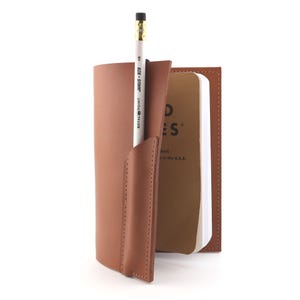May include: A brown leather notebook cover with a pencil holder, holding a white pencil with gold accents. A small notebook with the word "NOTES" is visible. The cover is made of stitched leather.