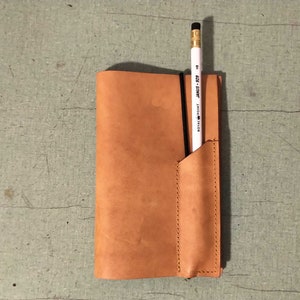 May include: A brown leather notebook cover with a pencil holder and a black elastic band. The pencil is white with black writing that says "STAEDTLER MARS 207".