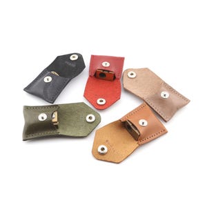 May include: Six small leather cases in black, red, olive green, tan, and brown. Each case is open, displaying a small, metallic object. The cases are secured with a snap closure. The cases are likely designed to hold small items.