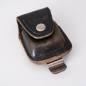 May include: A black leather case with a silver snap closure and the word "Zippo" embossed on the front. The case has a metal clip on the bottom.
