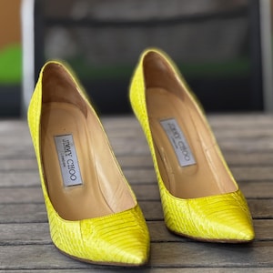 May include: A pair of vibrant yellow high-heeled pumps with pointed toes. The shoes feature a textured, snakeskin-like pattern and are labelled "JIMMY CHOO LONDON" inside. They are placed on a wooden surface.