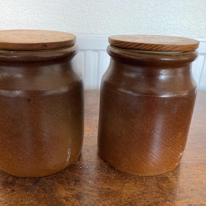 Könnte beinhalten: Zwei braune Keramikgläser mit Holzdeckeln. Die Gläser haben eine zylindrische Form mit einem etwas breiteren Boden und einem schmaleren Hals. Die Holzdeckel sind kreisförmig und passen gut oben drauf. Die Gläser stehen auf einer Holzoberfläche.