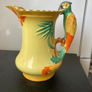 Pode incluir: Jarro de cerâmica amarelo com uma pega em forma de papagaio. O jarro apresenta detalhes pintados em laranja e verde, incluindo uma pega de papagaio, desenhos florais e de folhas. O jarro tem um bico largo e uma base redonda.