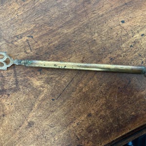 Könnte beinhalten: Eine dreizackige Gabel aus Messing mit einem langen, dünnen Stiel. Die Gabel liegt auf einer Holzoberfläche.