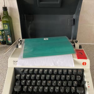 Puede incluir: Una máquina de escribir blanca con un teclado negro y una hoja de inserción verde. La máquina de escribir está etiquetada como "OLY! Moni".
