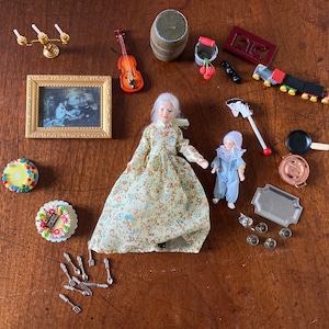 Puede incluir: Una escena de casa de muñecas en miniatura con una muñeca vestida con un vestido floral, una muñeca infantil con un mono azul, una imagen enmarcada, un tren de juguete, un pastel, un violín, un candelabro, un barril, un cubo, un caballo de balancín, una sartén, una bandeja y tazas de té.