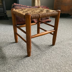 May include: A wooden stool with a woven seat. The stool has a simple design with four legs and a square seat.