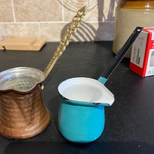 Può includere: Due piccole caffettiere. Una è color rame con un manico in ottone. L'altra è turchese con un manico nero. Entrambe le caffettiere hanno beccucci per versare. Le caffettiere sono su una superficie scura.