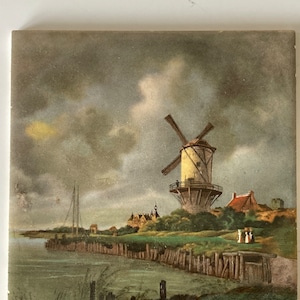 May include: A vintage tile depicting a Dutch windmill with a white body and brown blades. The windmill is situated on a grassy hill with a river in the foreground. The sky is cloudy and gray with a hint of yellow in the distance.