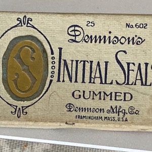 Op de afbeelding: Vintage doosje met Dennison's Initial Seals. De rechthoekige doos heeft een lichtbeige achtergrond. De tekst "Dennison's Initial Seals" staat in donkerblauwe letters. De doos heeft een gouden initiaal "S" in een blauw en goud ovaal.