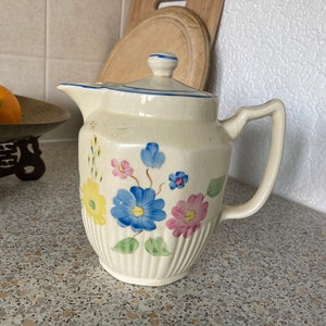 Può includere: Una teiera in ceramica bianca con un design floreale. La teiera ha un motivo floreale blu, giallo e rosa con foglie verdi. La teiera ha un coperchio e un manico.