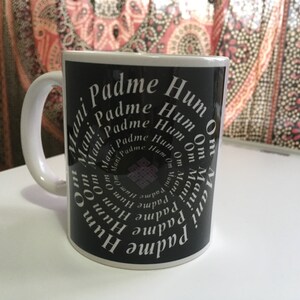 Puede incluir: Una taza de cerámica blanca con fondo negro con el texto "Om Mani Padme Hum" en espiral. La taza tiene un asa blanca y un borde blanco. El diseño incluye un símbolo de nudo central.