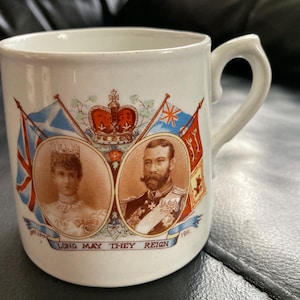 Op de afbeelding: Witte keramische mok met een handvat, versierd met portretten van een man en een vrouw, vlaggen en een kroon. De mok heeft de tekst "LONG MAY THEY REIGN". De portretten staan in ovale kaders.