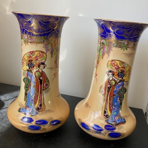 Peut inclure: Deux vases décoratifs en céramique avec une base beige et des accents bleus et dorés. Chaque vase présente une scène peinte d'une femme avec un parapluie. Les vases ont un sommet évasé et une base bulbeuse.