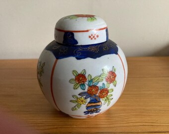 Peut inclure: Petit pot en céramique rond et couvert, à base blanche et bord bleu. Le pot est orné de motifs floraux orange et jaunes et d'accents rouges. Le couvercle présente un motif floral et des points rouges.