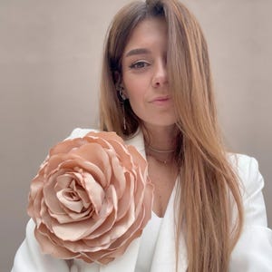 May include: A large, peach-colored fabric rose is attached to a white blazer. The rose is the focal point, with multiple layers of petals. The blazer has a classic cut and is worn over a white top. The background is a neutral color.