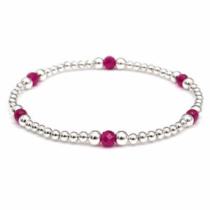 May include: A stretch bracelet composed of small, polished silver-toned spherical beads interspersed with five vibrant, faceted magenta-coloured round beads. The design features a repeating pattern of metallic silver spheres and bright pink accents.
