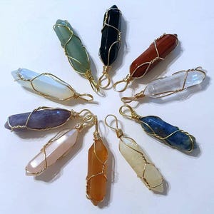 May include: A selection of gemstone pendants in diverse colours, such as green, black, red, and blue. Each crystal is wrapped in gold-coloured wire with a loop for hanging. The pendants are arranged in a circular pattern.