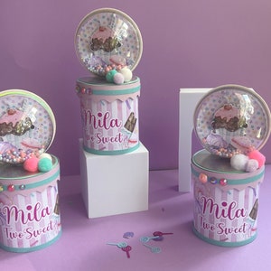 May include: Three pink and white striped favour boxes with clear lids. Each box has a cupcake design with sprinkles and a pink cherry on top. The boxes are decorated with pink and white pom-poms and rhinestones. The boxes are labelled "Mila Two Sweet".
