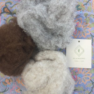 May include: Three piles of wool fiber in different colors: light gray, brown, and beige. The wool is fluffy and soft, and it is arranged on a patterned fabric background. A white card with the text "Plainsong Farm & Fiber" is visible in the upper right corner of the image.