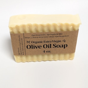 May include: A rectangular bar of off-white soap with a wavy top edge, wrapped in a brown paper label. The label reads: Organic Extra Virgin Olive Oil Soap, 4 oz., OpeSoapsLLC, Springfield MO, and lists ingredients as organic extra virgin olive oil, water, sodium hydroxide.