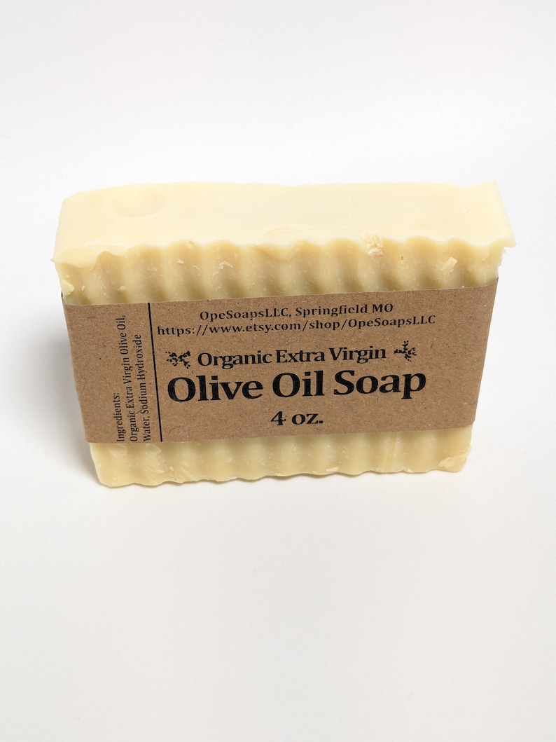 May include: A rectangular bar of off-white soap with a wavy top edge, wrapped in a brown paper label. The label reads: Organic Extra Virgin Olive Oil Soap, 4 oz., OpeSoapsLLC, Springfield MO, and lists ingredients as organic extra virgin olive oil, water, sodium hydroxide.