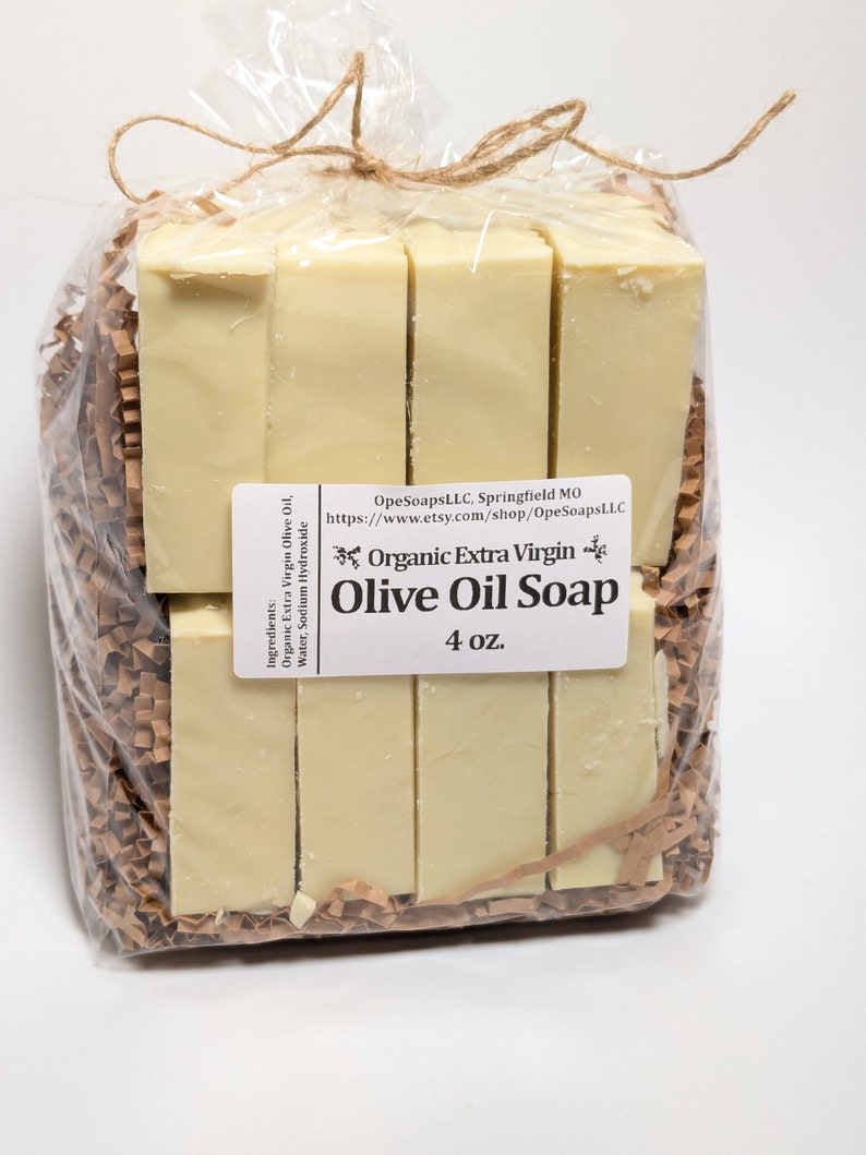 May include: A package of six bars of organic extra virgin olive oil soap, wrapped in clear plastic and brown paper. The label reads "Organic Extra Virgin Olive Oil Soap 4 oz."