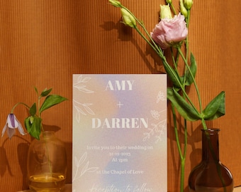 May include: A wedding invitation with a holographic iridescent background and white text. The invitation reads "AMY + DARREN Invite you to their wedding on 21-12-2023 At 2pm at the Chapel of Love Reception to follow."