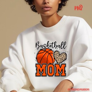 May include: White sweatshirt with the text "Basketball Mom" in black and orange. A basketball and a leopard print heart are above the text.