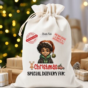Puede incluir: Una bolsa de regalo de Navidad de lona blanca con un sello de correo de Santa rojo y blanco, un sello de Polo Norte y un sello de No abrir hasta el 25 de diciembre. La bolsa presenta una ilustración de dibujos animados de un niño pequeño con un gorro de Papá Noel y una chaqueta verde. El texto "Christmas Special Delivery For:" está impreso en la bolsa.