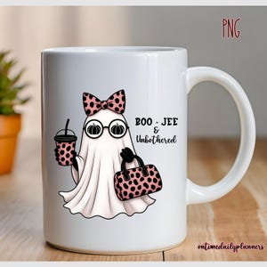 May include: White ceramic mug featuring a cartoon ghost with a pink bow, glasses, and a purse. The ghost holds a coffee cup and is labeled "BOO - JEE & Unbothered". The mug has a handle and is on a wooden surface.