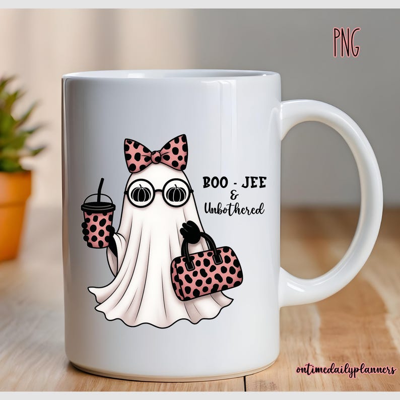 May include: White ceramic mug featuring a cartoon ghost with a pink bow, glasses, and a purse. The ghost holds a coffee cup. Text on the mug reads "BOO - JEE & Unbothered". The mug has a handle and is on a wooden surface.