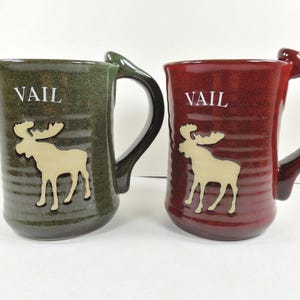 May include: Two ceramic mugs, one green and one burgundy, with large handles. Each mug features the word "VAIL" in white and a tan moose silhouette. The mugs are likely for beverages.