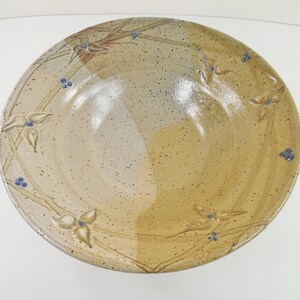 May include: A round ceramic bowl with a speckled beige and tan glaze. The interior features a raised relief pattern of slender stems, three-petaled flowers, and small clusters of blue berries arranged in a circular design around the rim.