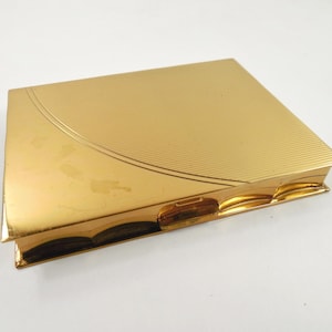 May include: A rectangular gold-toned metal compact case with a polished finish. The top surface features a curved, etched line pattern on one side, while the other side remains smooth. The case includes a small, centered latch mechanism on the front edge.
