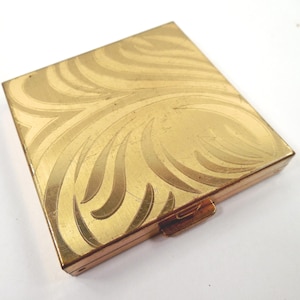 May include: A square gold-tone metal compact case featuring an engraved swirling leaf pattern on the top lid. The compact has a hinged closure mechanism on the side and a polished metallic finish with visible fine surface scratches.
