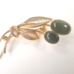 May include: A gold-toned brooch featuring a filigree design with two oval, green gemstones. The brooch has a leaf and stem design, with the gemstones set in a gold-colored frame. The filigree work adds a delicate touch to the jewelry.