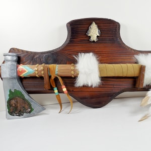 May include: A decorative Native American-style axe mounted on a wooden plaque. The axe head is silver with a bear carving, and the handle is adorned with leather, fur, and feathers. The plaque is dark brown with a carved arrowhead.